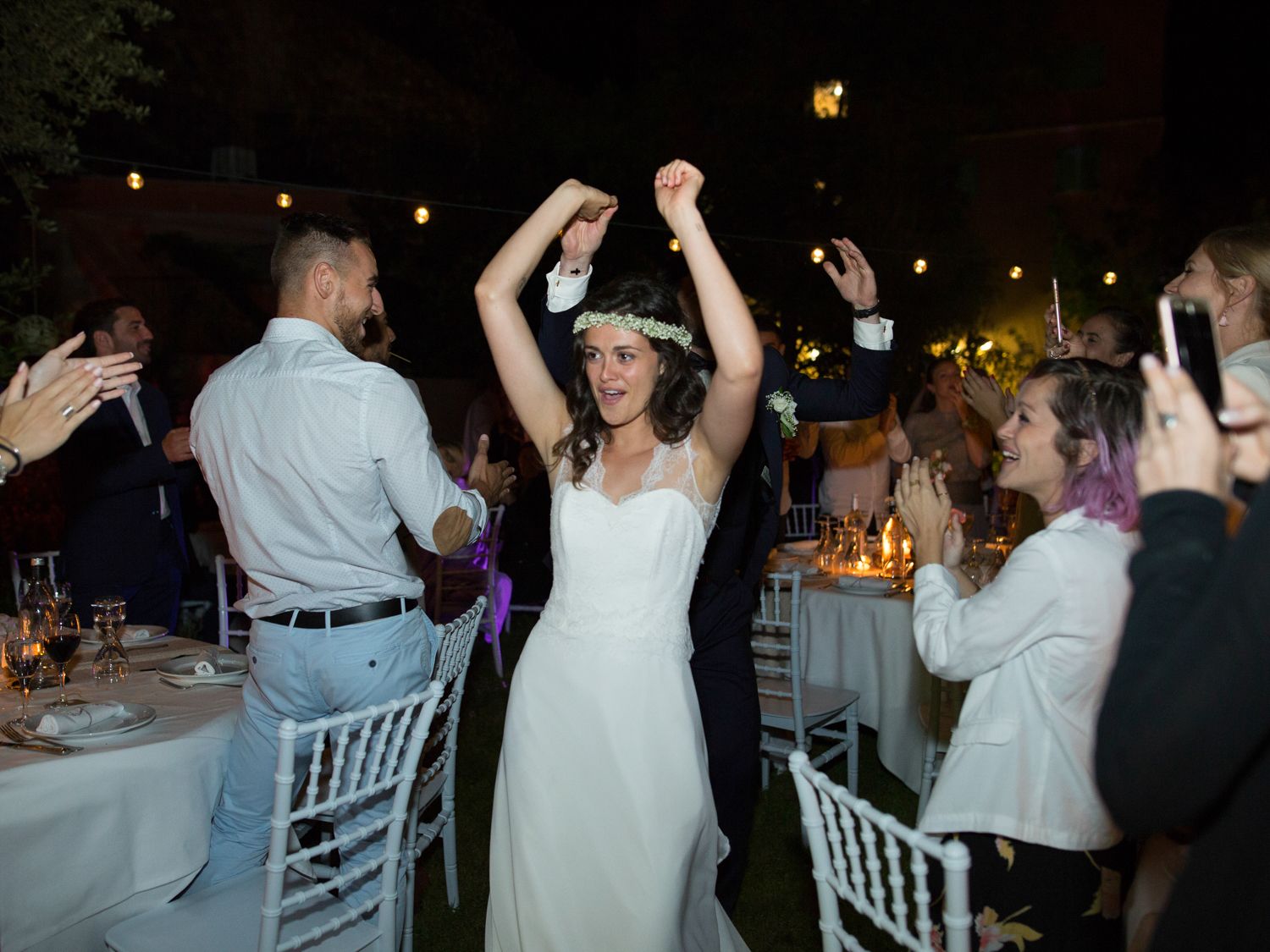 La mariée en robe blanche lève les mains avec joie tandis que les invités célèbrent une élégante réception de mariage en plein air