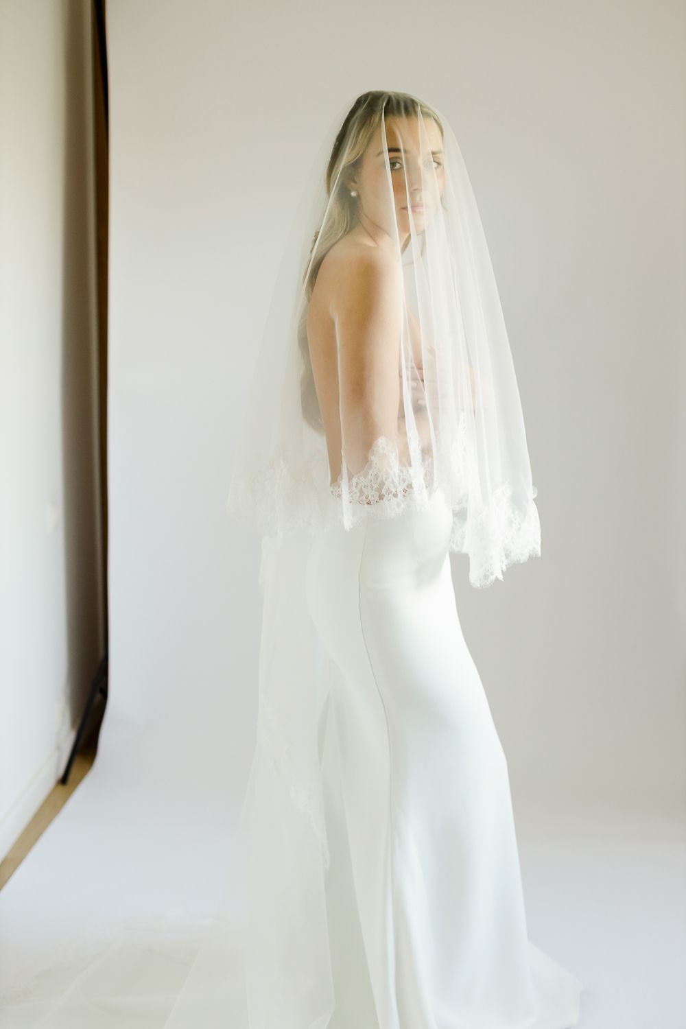 Bride in a sleek white gown and lace-trimmed veil draped over her face, posing elegantly in a bright studio.