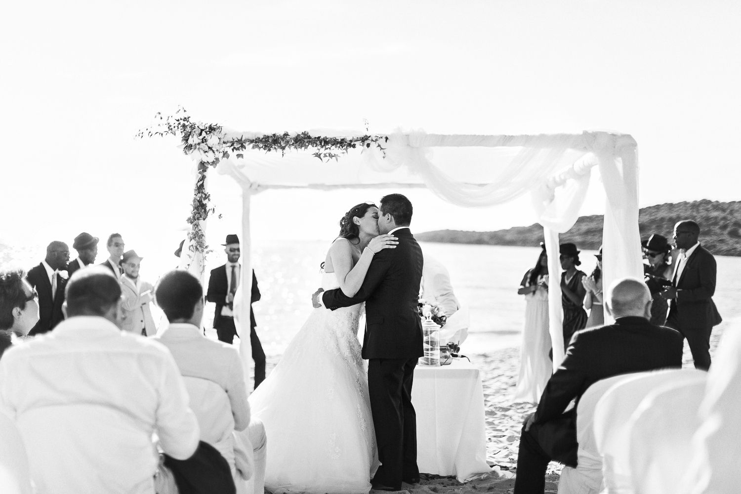 Cérémonie de mariage en noir et blanc sur la plage : les mariés s’embrassent sous une arche fleurie, les invités sont assis sur des chaises