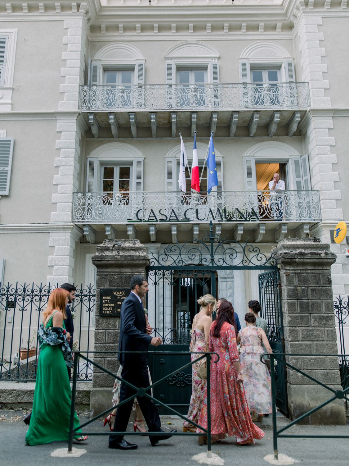 Elegantly dressed wedding guests arriving at Casa Comunale, a grand white Italian villa with ornate balcony and flags.