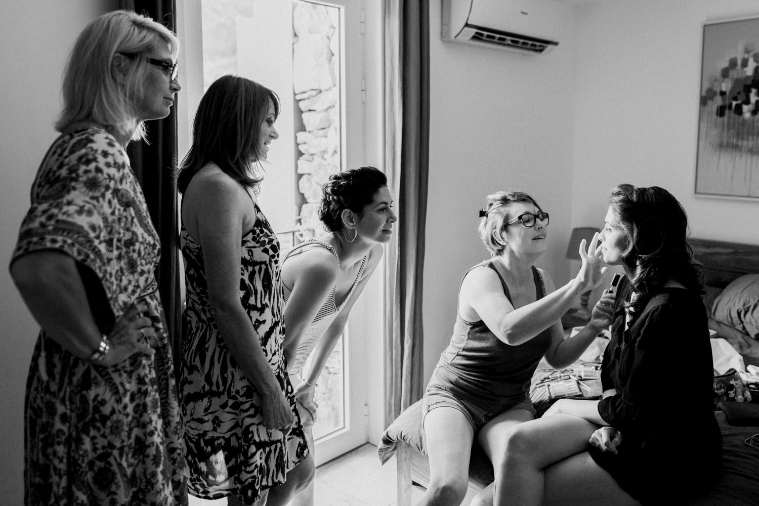 Black and white photo of bridesmaids helping bride with makeup and hair before wedding ceremony indoors.
