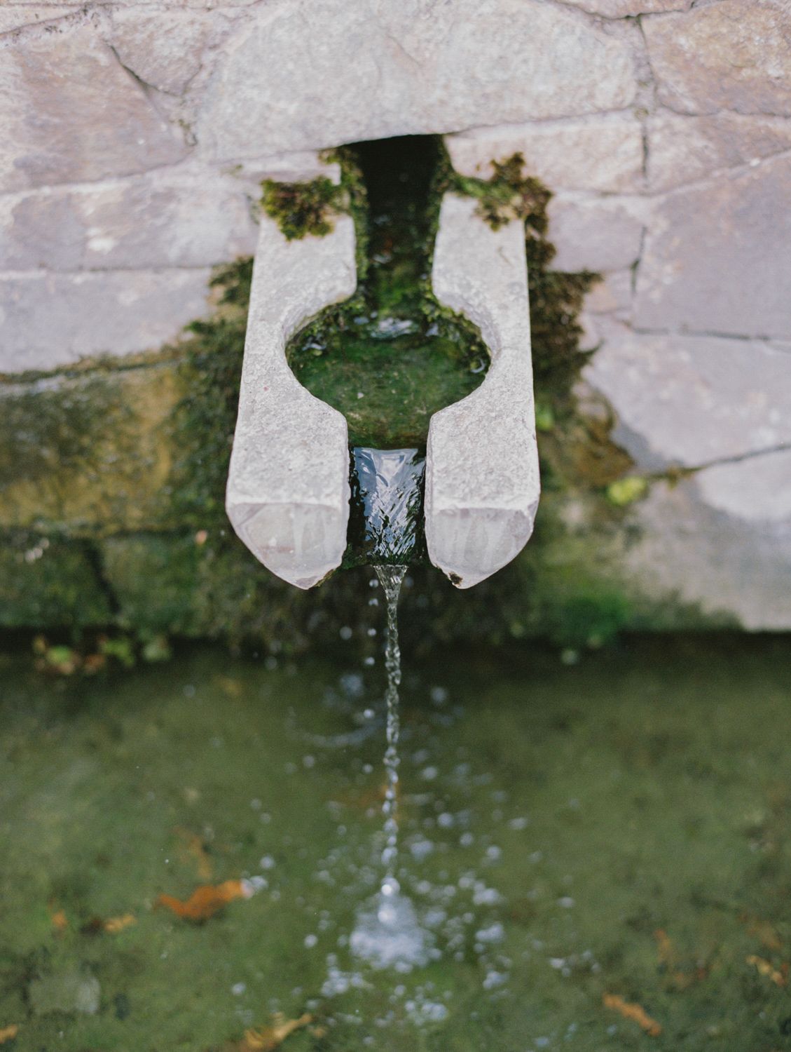 Water flowing from a stone fountain spout into a mossy basin, set against an ancient weathered stone wall.