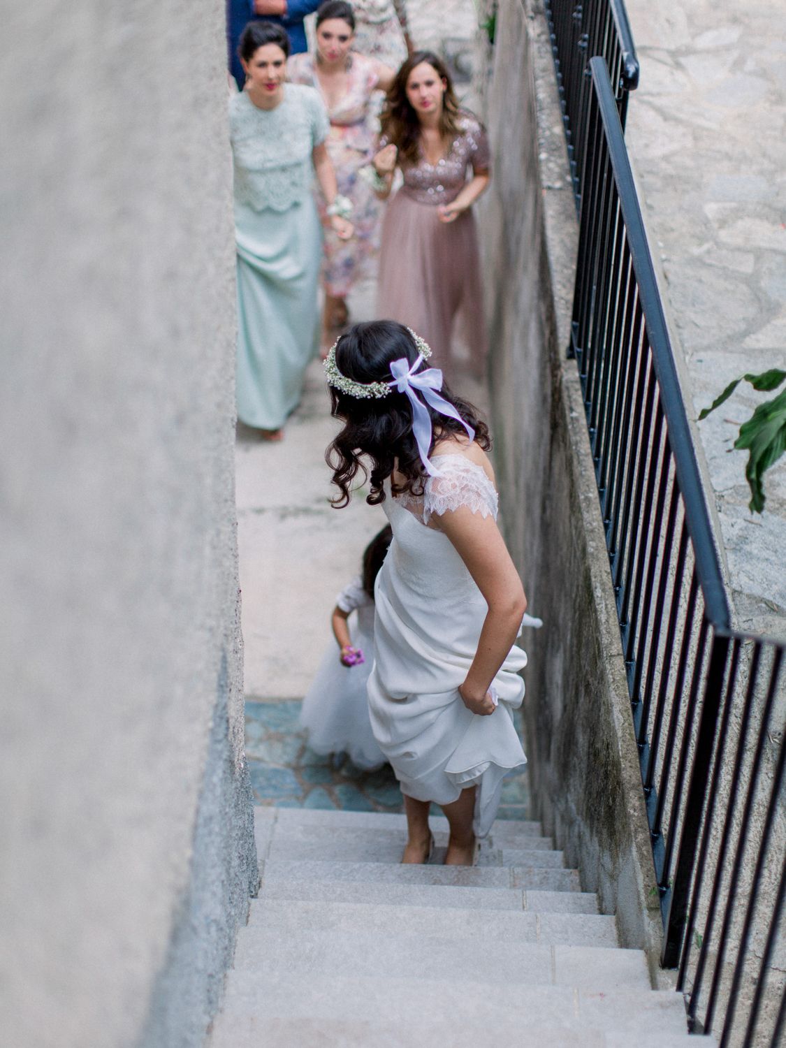 A flower girl in a white dress and floral crown walks up stone stairs ahead of bridesmaids in mint and mauve gowns.
