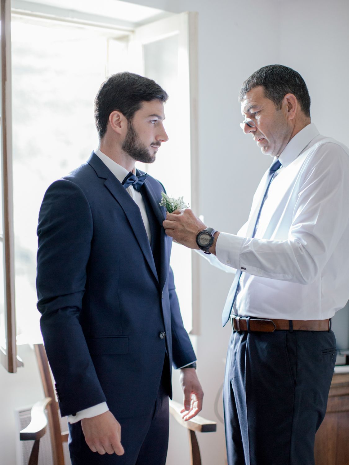 Older man pinning boutonniere on groom in navy suit before wedding ceremony indoors.