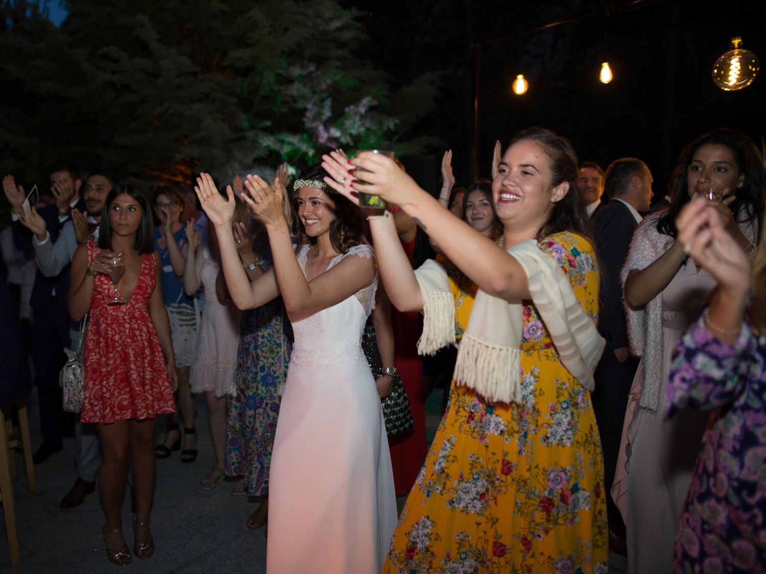 Des invités dansent et applaudissent lors d'une réception de mariage en plein air, sous des guirlandes lumineuses.