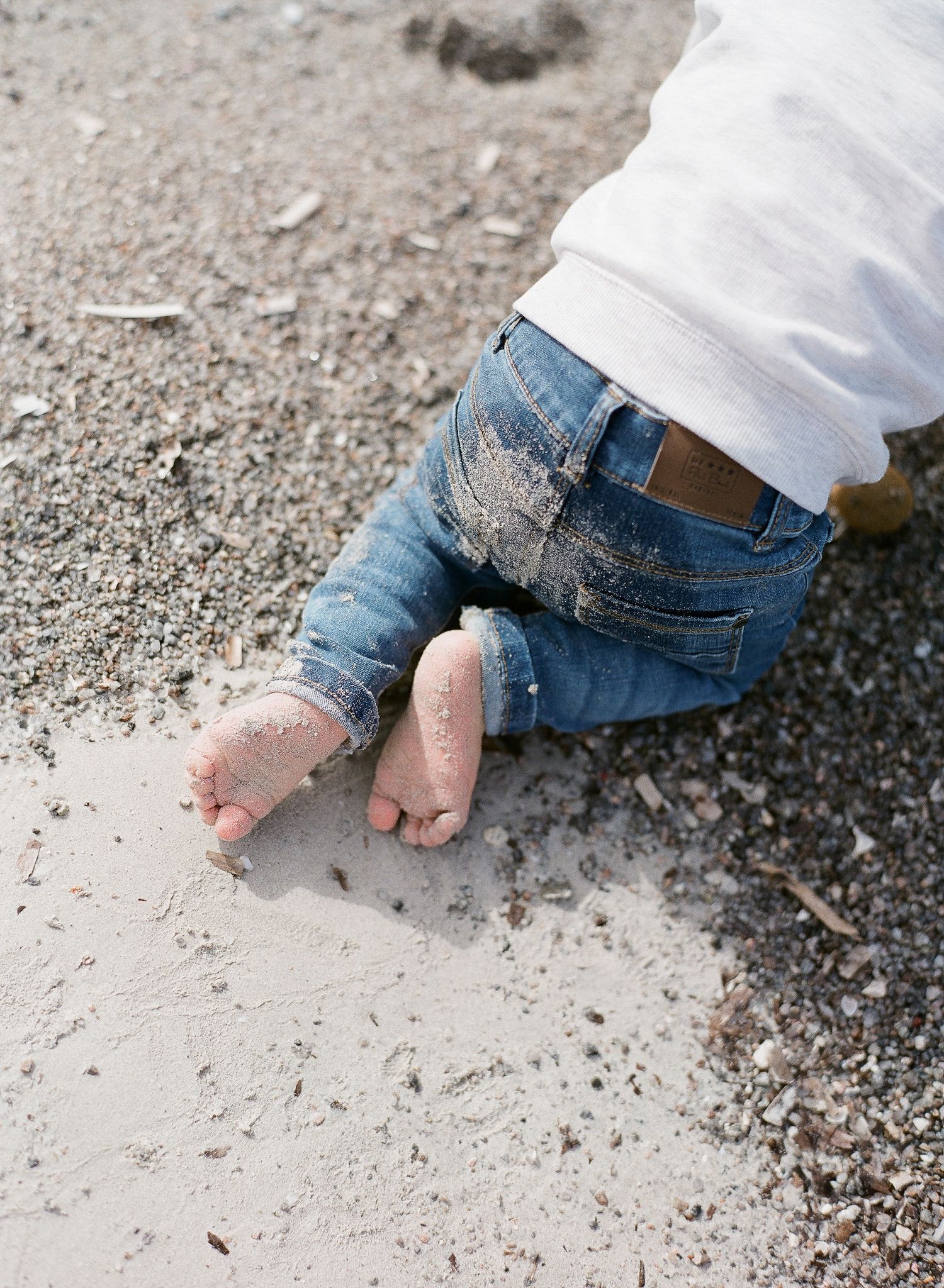 Gros plan sur les pieds et les mains sablonneux d'un bébé en jean rampant sur la plage de Santa Giulia.