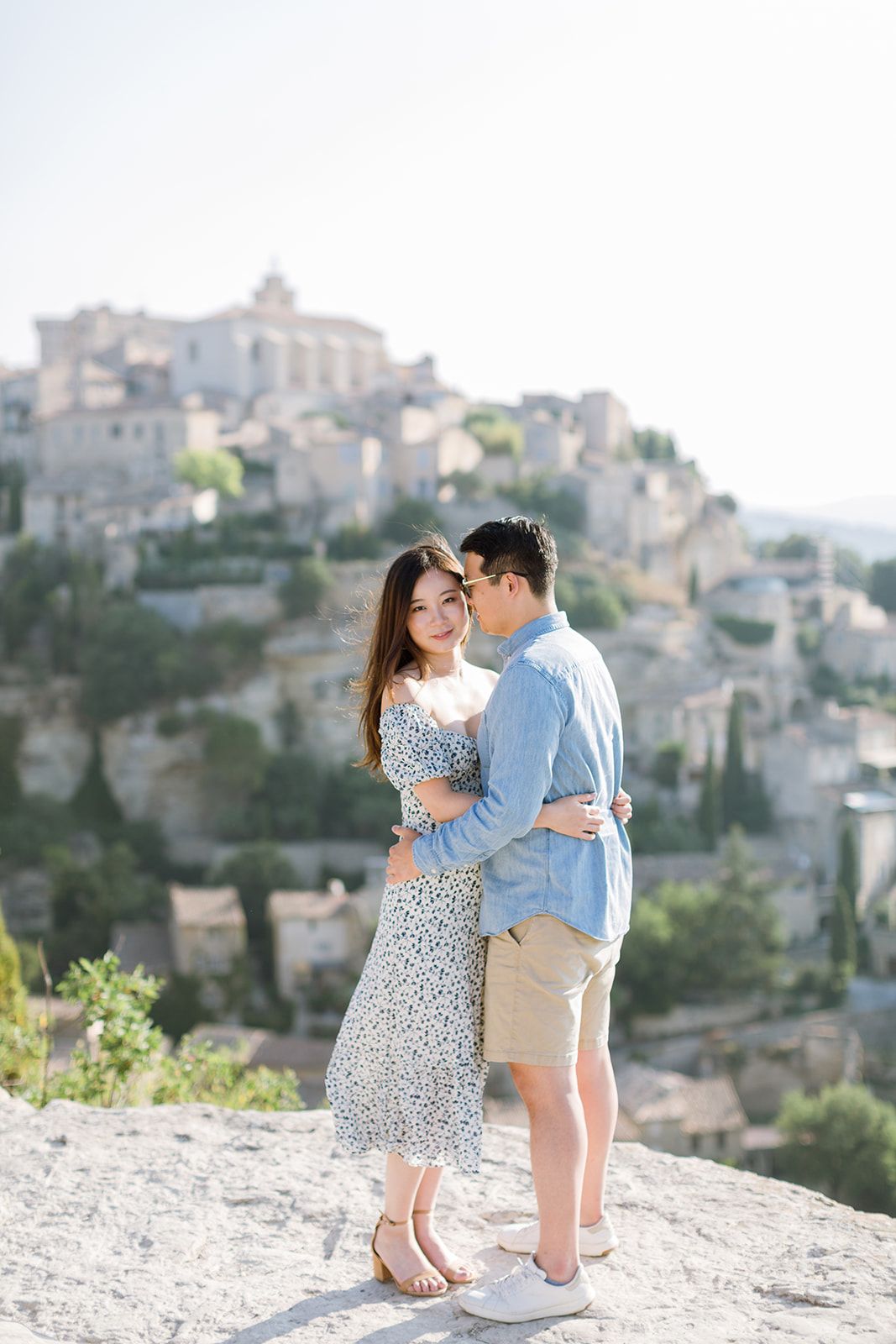 Un couple s'enlace amoureusement sur un promontoire rocheux baigné de soleil surplombant le village médiéval de Gordes, en Provence.