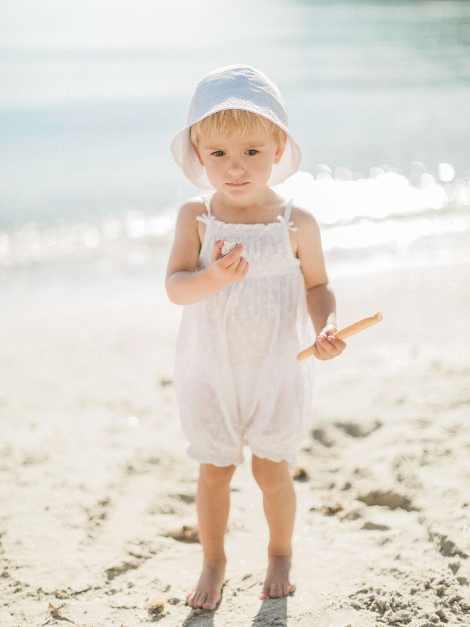 Adorable bambin en barboteuse blanche et chapeau de soleil, pieds nus sur une plage de sable ensoleillée.