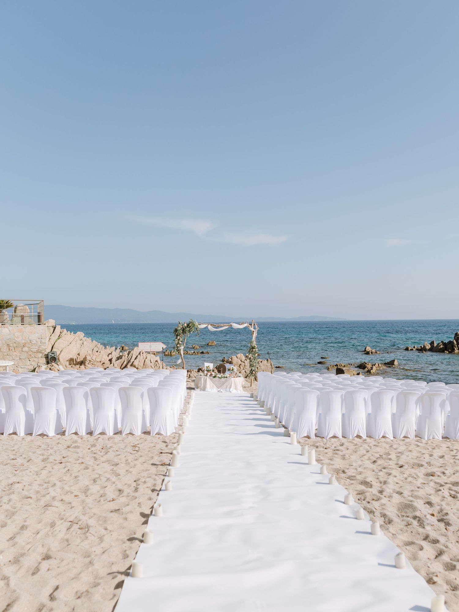 Installation élégante pour une cérémonie de mariage sur la plage, avec des housses de chaise blanches et une allée menant à une arche fleuri