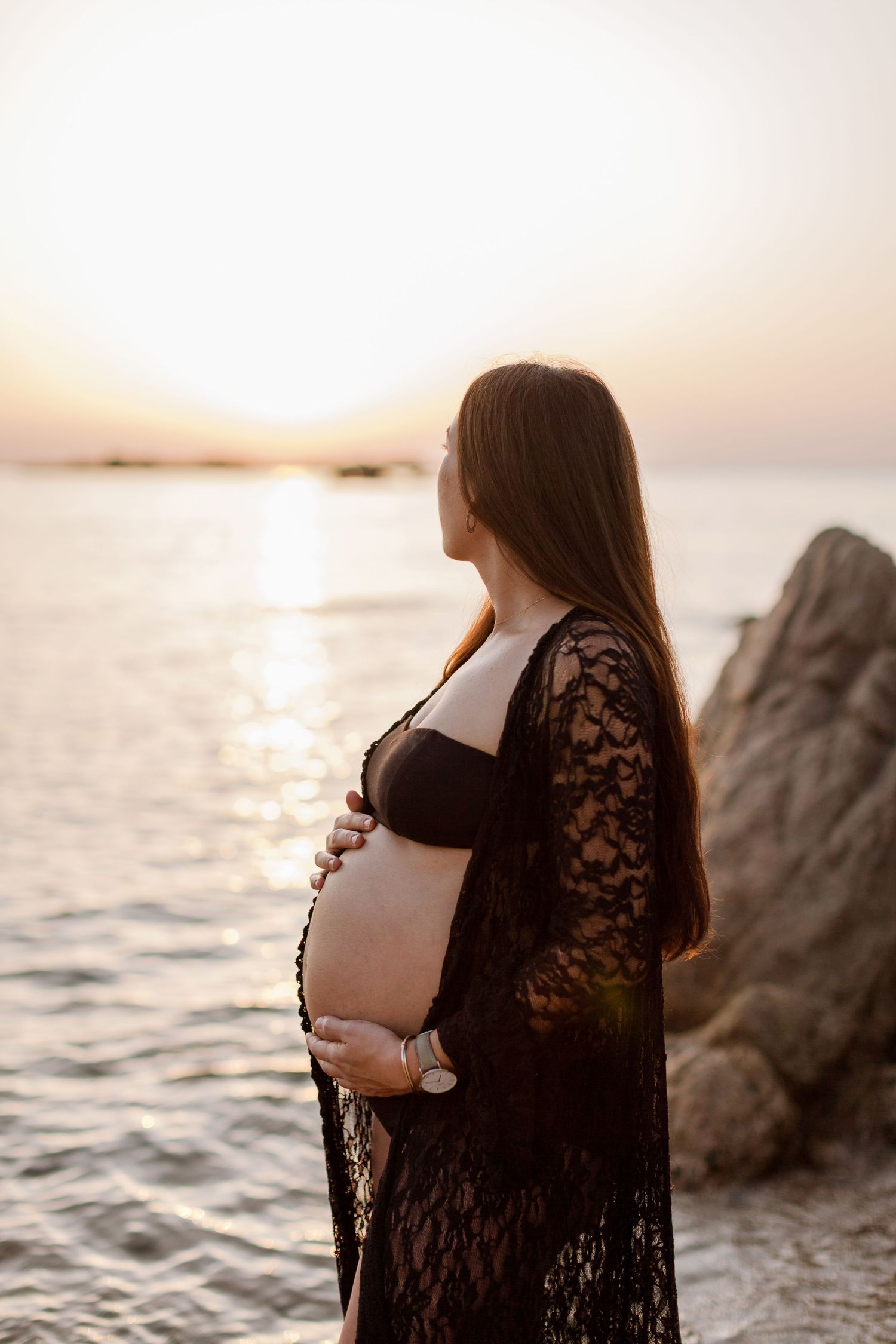 Profil latéral d'une femme enceinte vêtue d'une robe en dentelle noire, se tenant le ventre au coucher du soleil à Bonifacio, Corse.