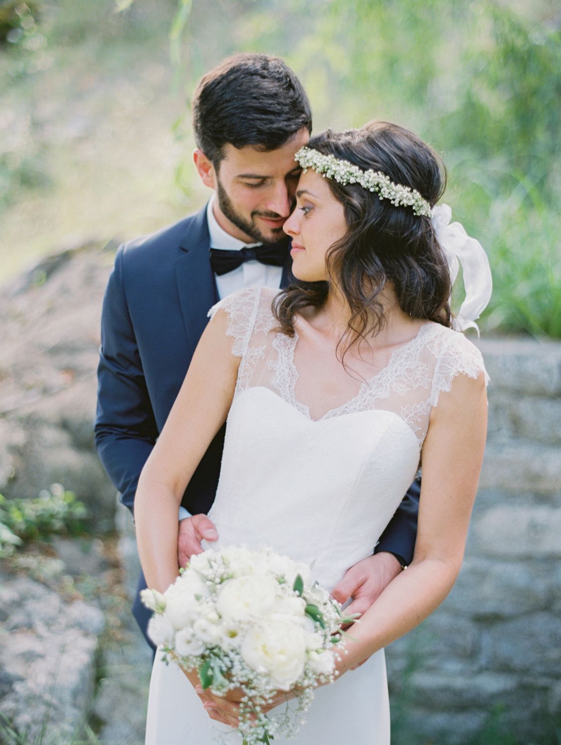 Bride in lace gown with floral crown holds white bouquet as groom in navy suit embraces her outdoors.