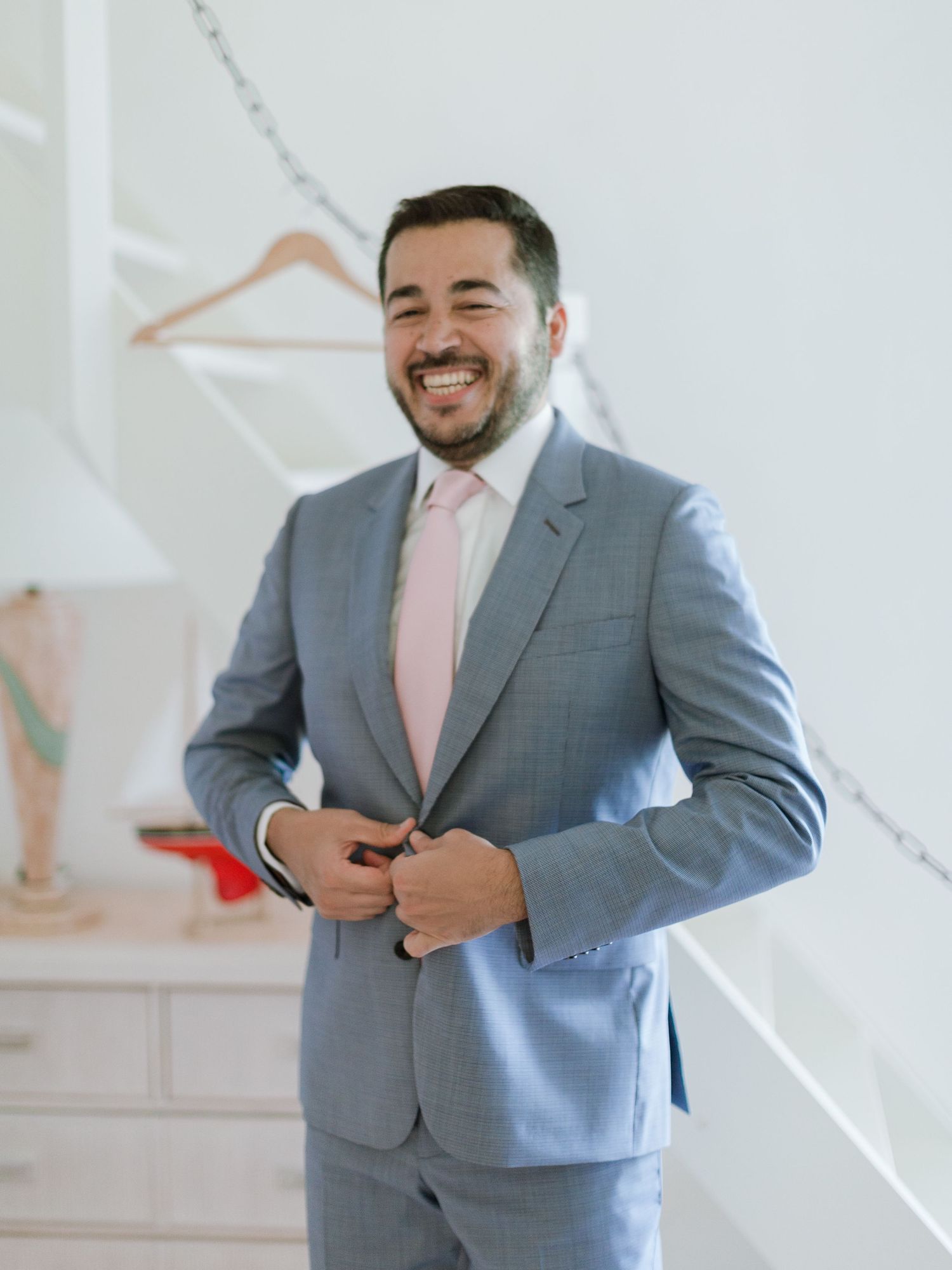 Un homme souriant, vêtu d'un costume bleu clair et d'une cravate rose, boutonne sa veste dans une pièce d'un blanc éclatant.