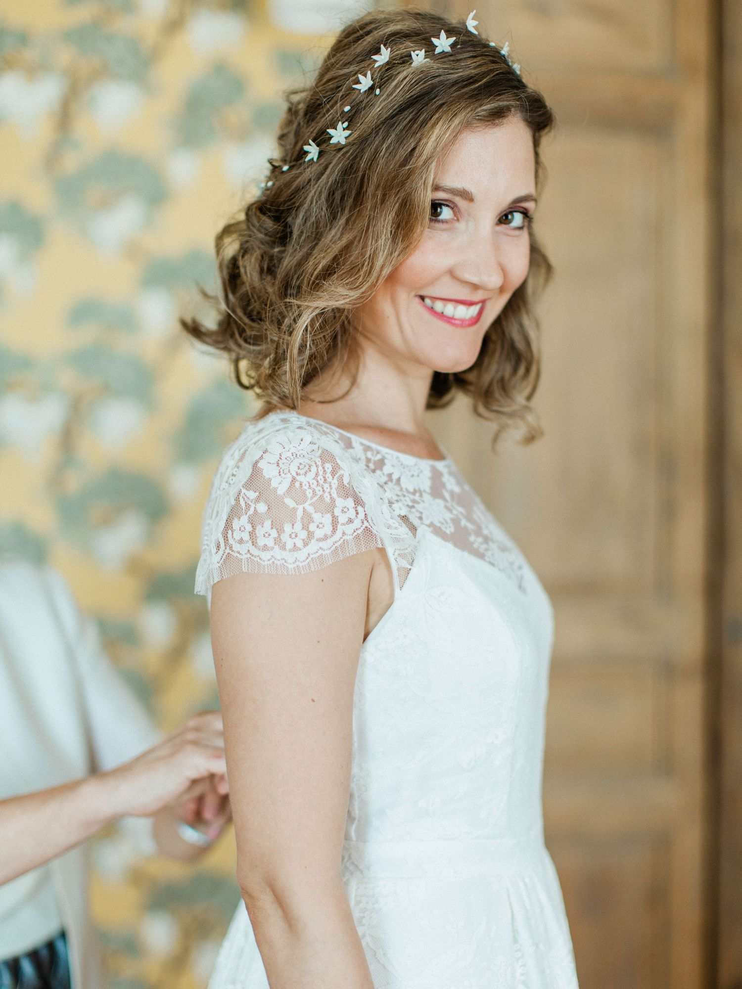 Smiling bride in lace cap sleeve wedding dress with star hair crown, short wavy updo, being helped with dress buttons.