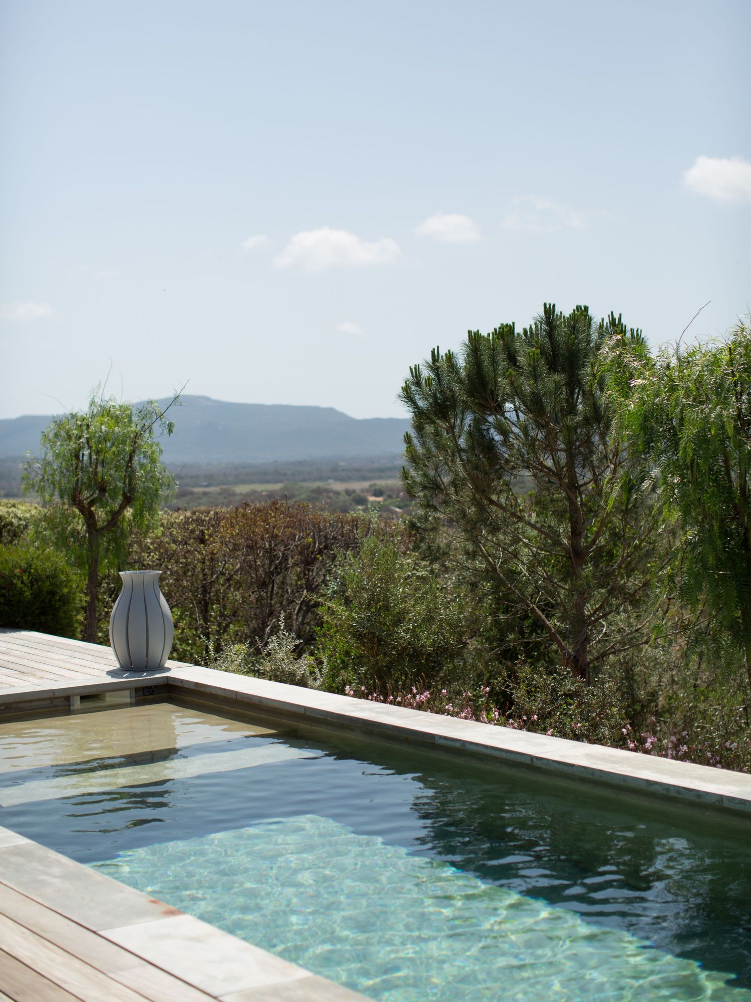 Piscine à débordement avec terrasse en bois, vase en céramique blanche, entourée d'un paysage méditerranéen et de vues sur les montagnes.