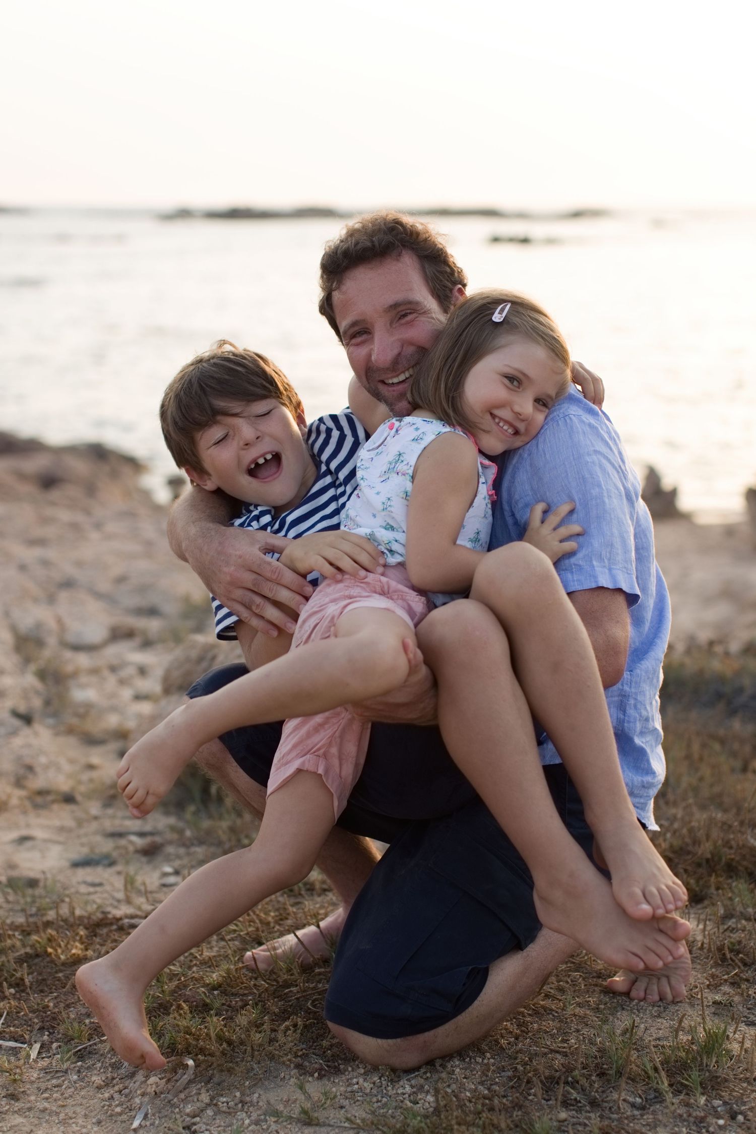 Un père heureux, assis sur le sable de la plage, rit et enlace ses deux jeunes enfants au coucher du soleil à Bonifacio.