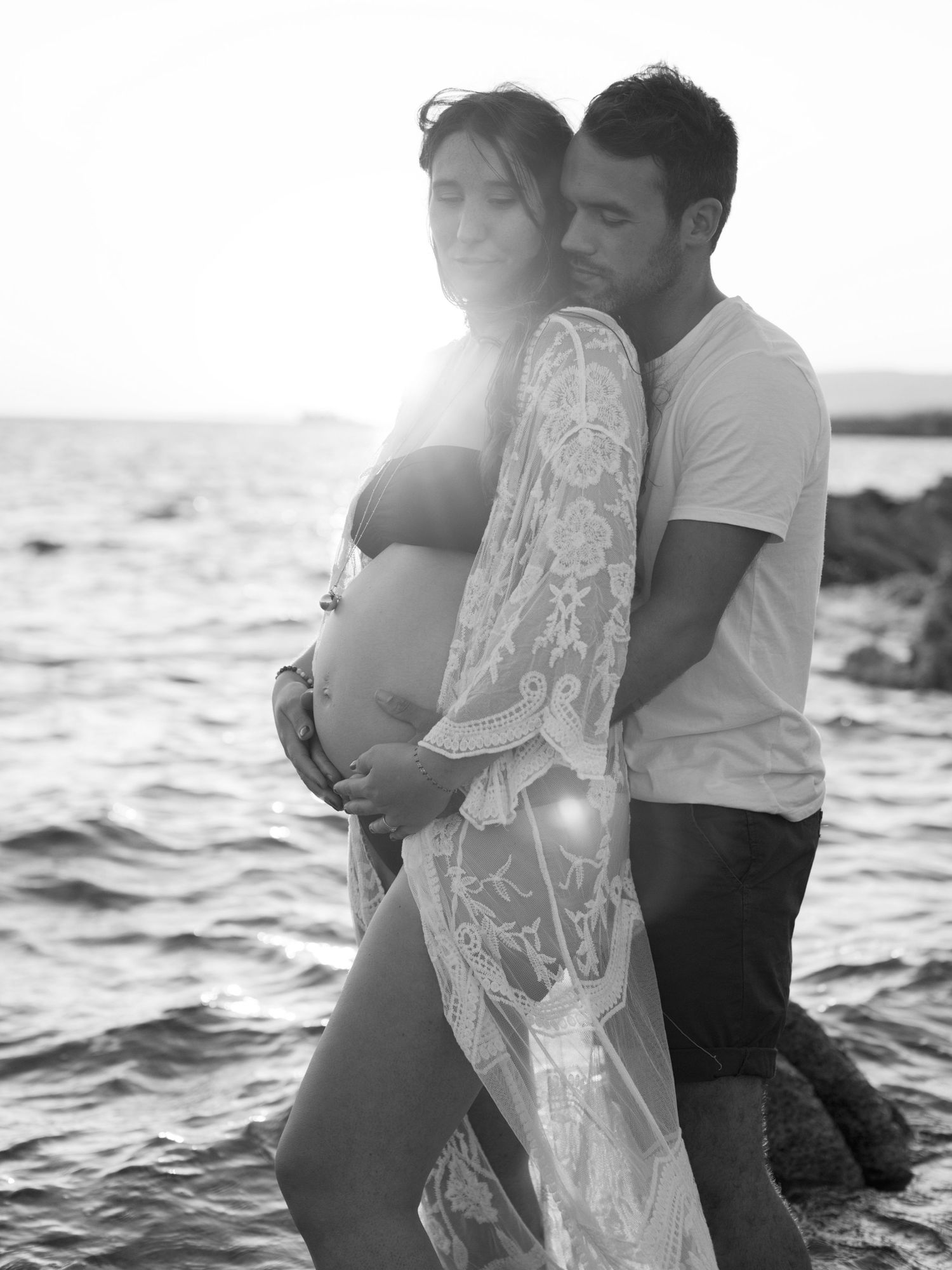 Black and white maternity photo of couple embracing by the sea at sunset.