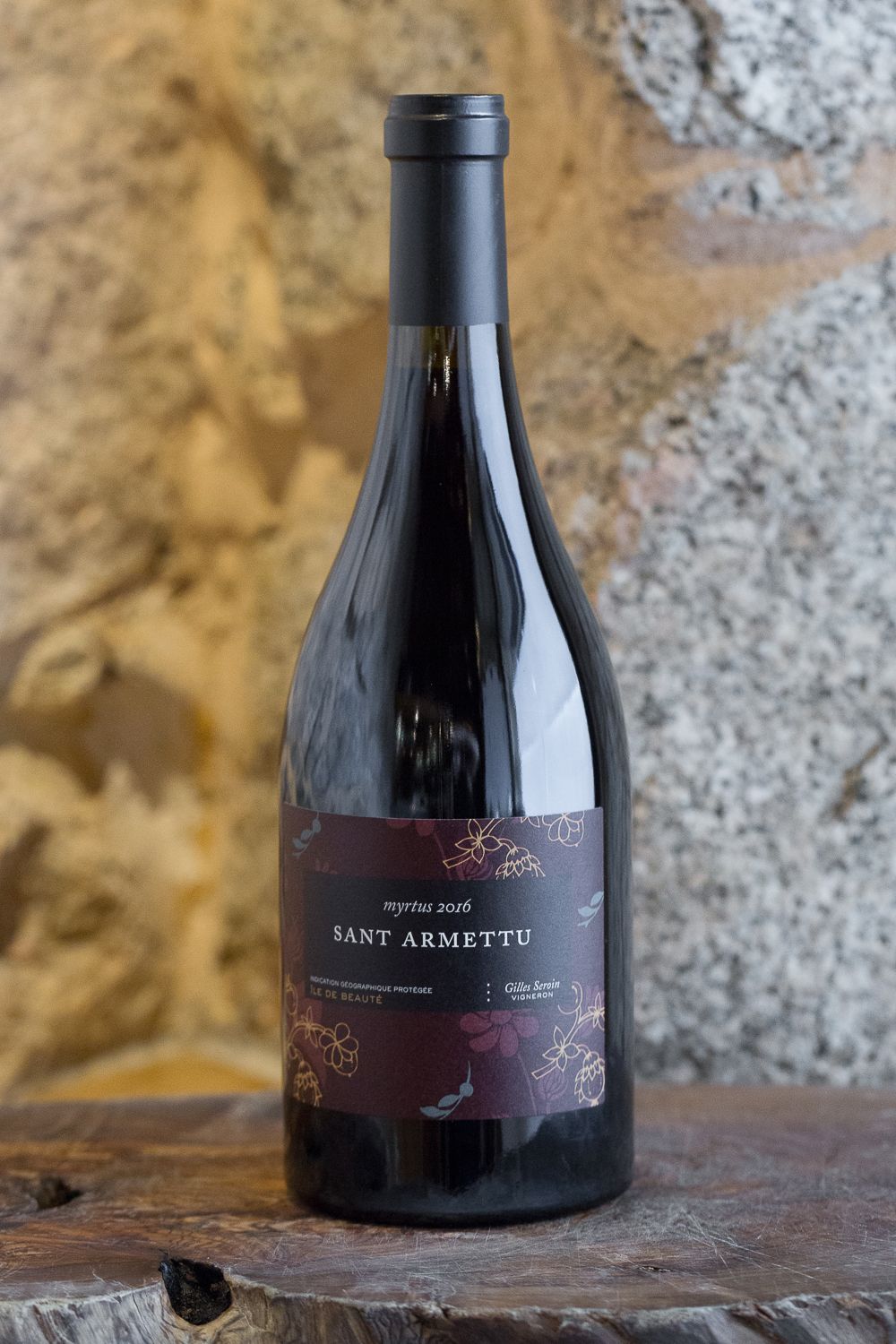 Bouteille de vin rouge Sant Armettu Myrtus 2016 sur une surface en bois avec un mur en pierre en arrière-plan.