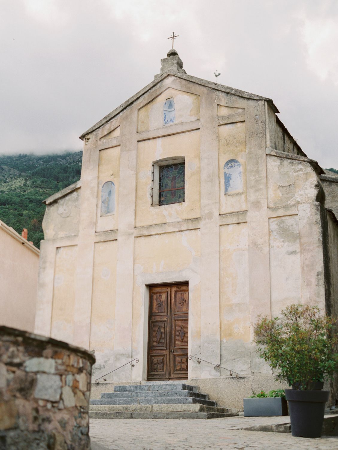 Historic yellow stone church with wooden doors, cross atop facade, stone steps, and lush green hills in background.