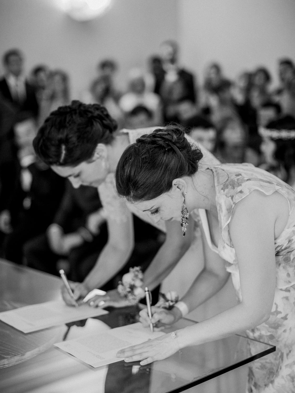 Black and white photo of a bride and groom signing wedding documents at a ceremony with guests seated in background.