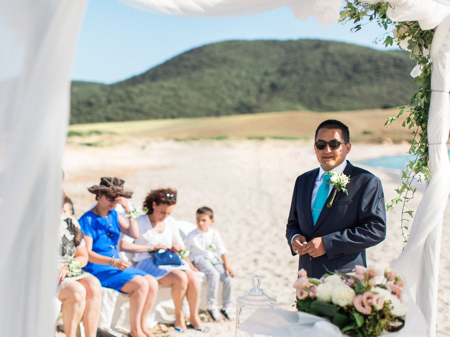 Le marié, vêtu d'un costume bleu marine et d'une cravate turquoise, se tient debout devant l'autel de la cérémonie sur la plage