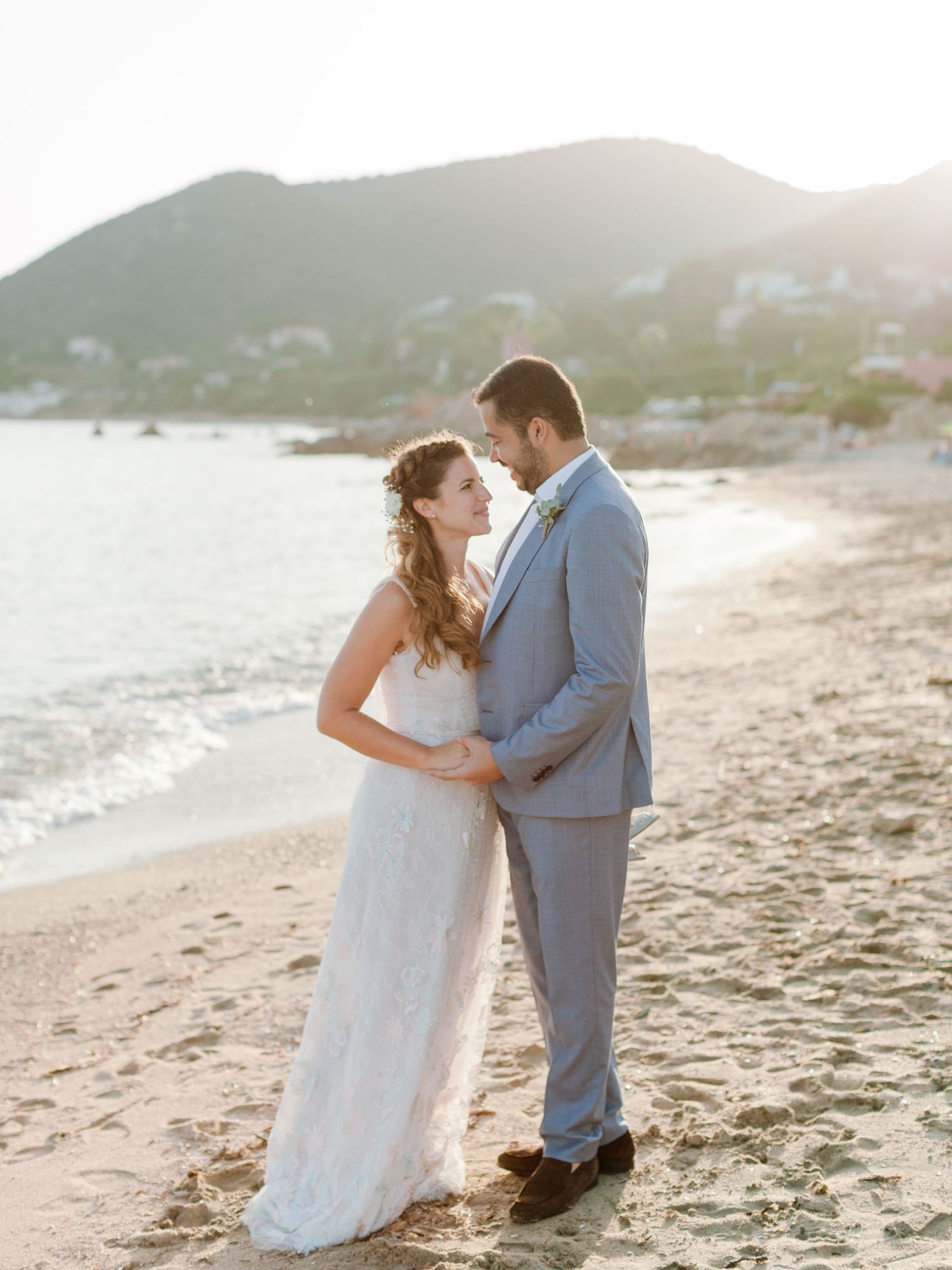 Les mariés partagent un moment romantique sur une plage de sable à l'heure dorée, avec l'océan et les montagnes en arrière-plan.