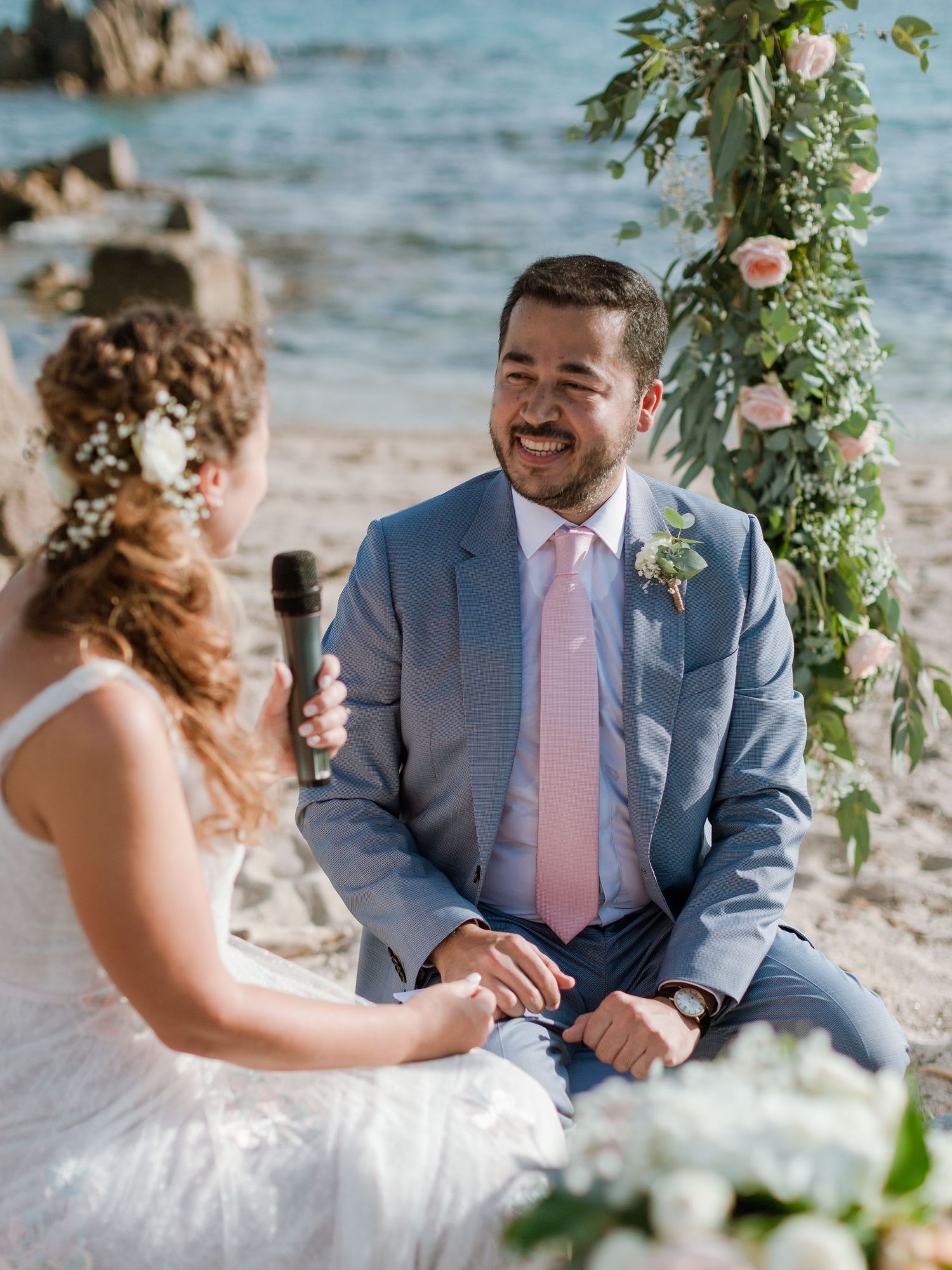 Marié en costume bleu et cravate rose riant pendant la cérémonie de mariage sur la plage, avec une arche florale en arrière-plan.