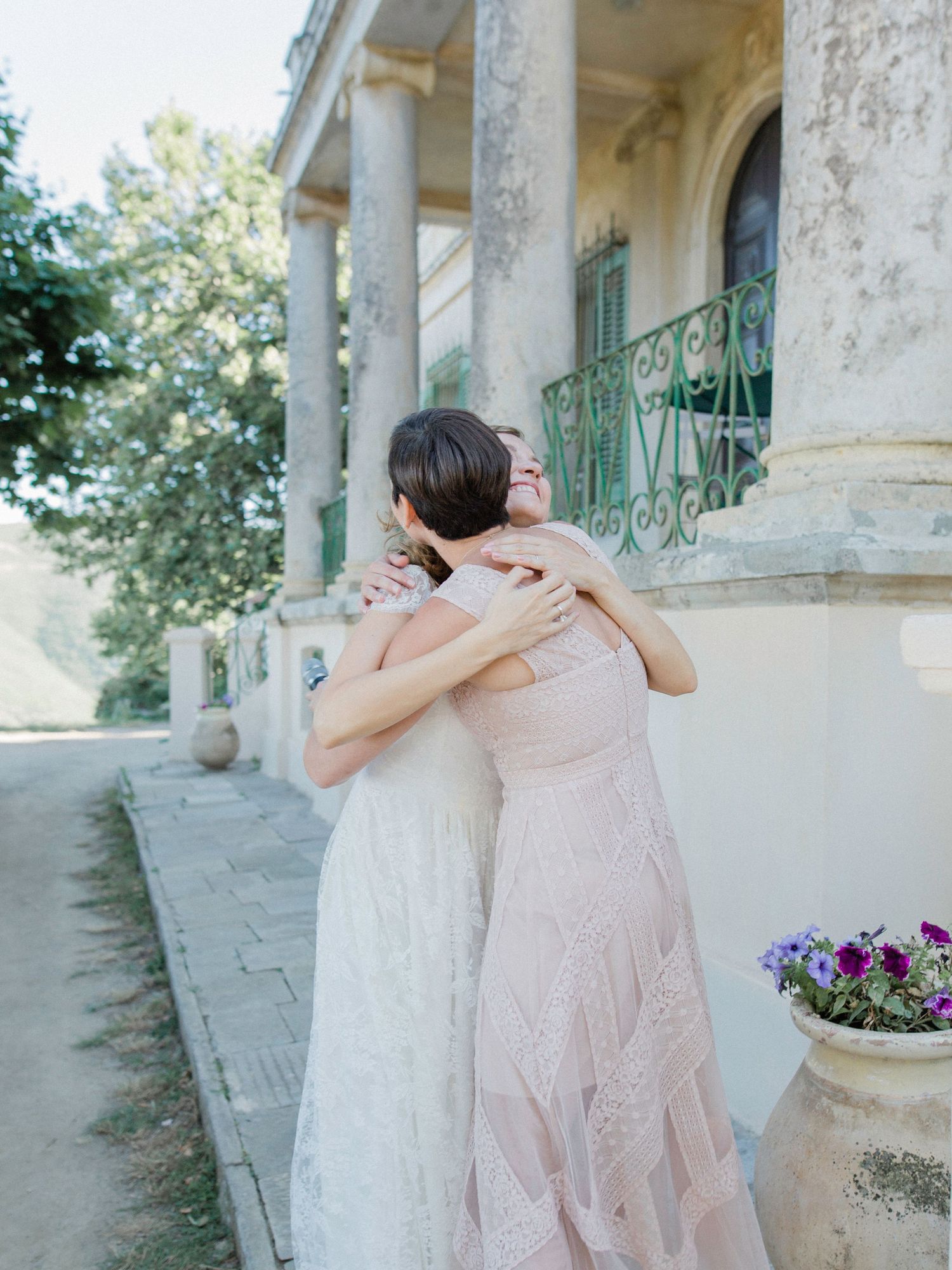 Two brides in elegant white and blush gowns share a heartfelt embrace outside a classic villa.