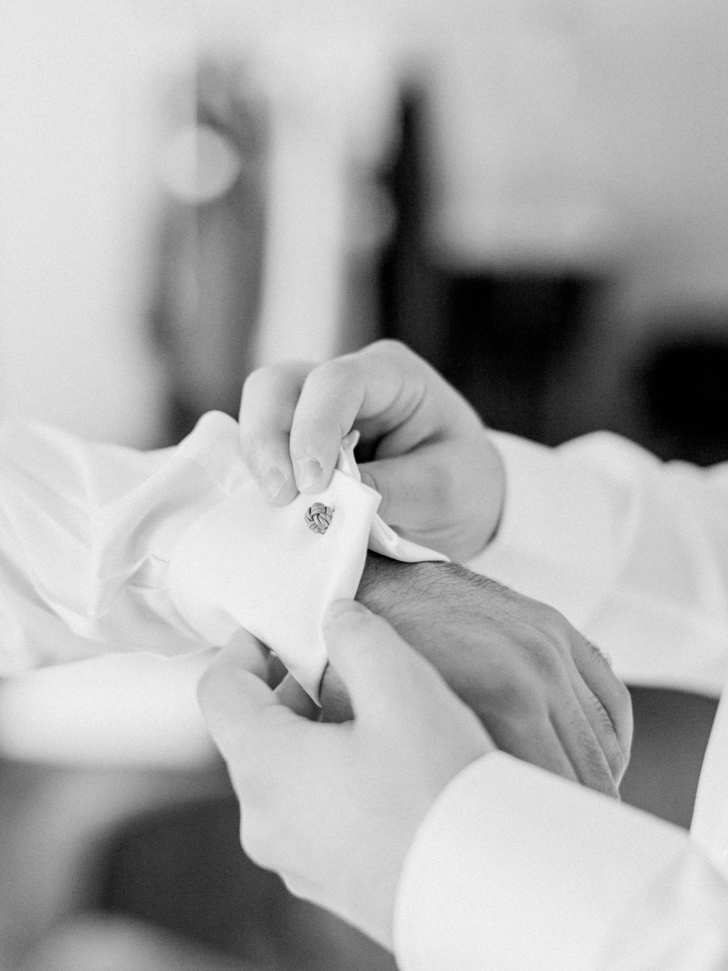 Black and white photo of a groom fastening cufflinks on his dress shirt sleeve before the wedding ceremony.