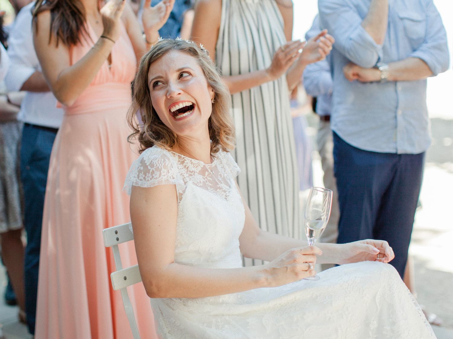 Joyful bride in white lace dress laughing while seated, surrounded by guests in pastel gowns at an outdoor wedding.