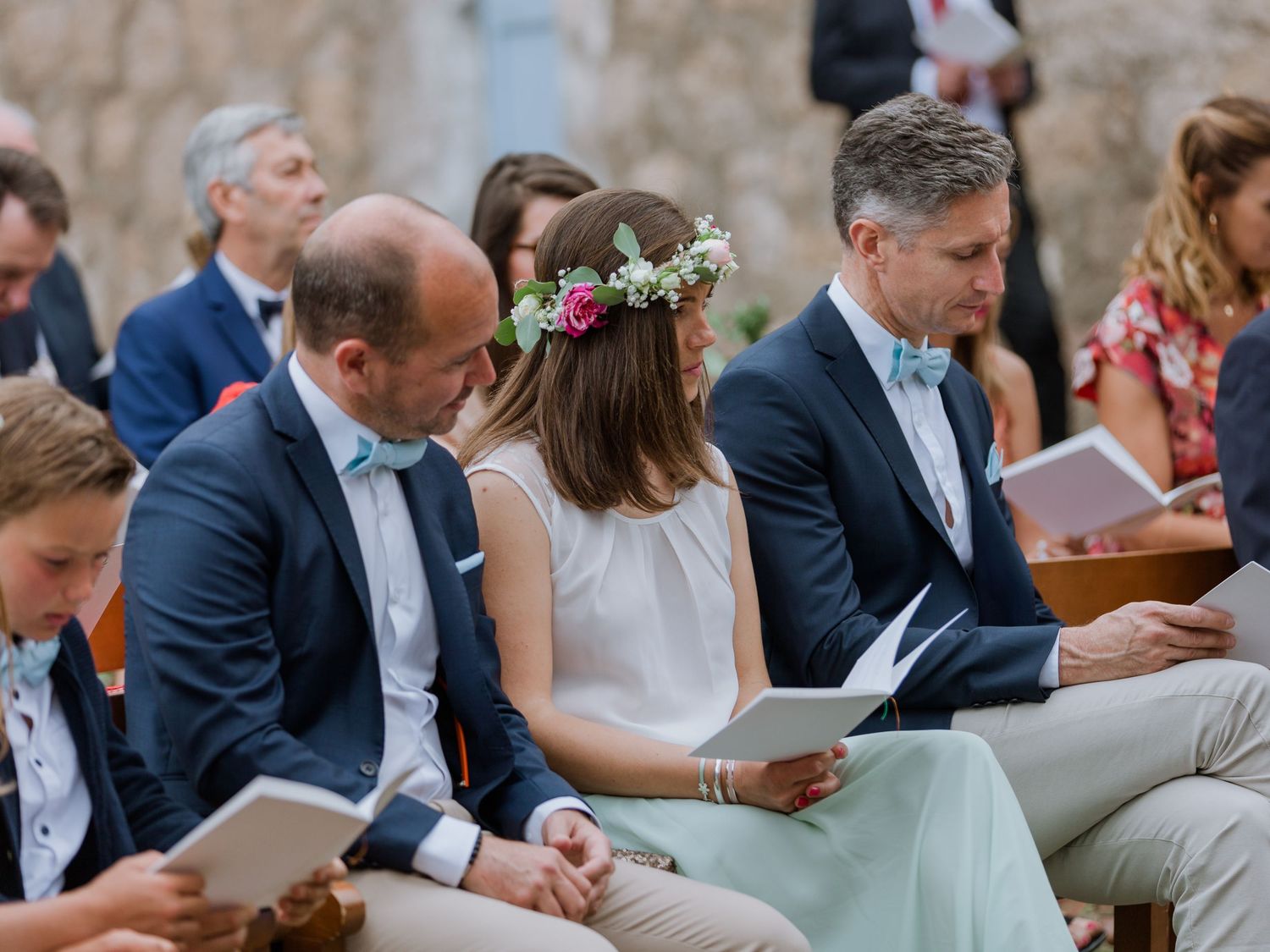 Des invités, assis à une cérémonie de mariage en plein air, lisent les programmes, une jeune fille portant une couronne de fleurs