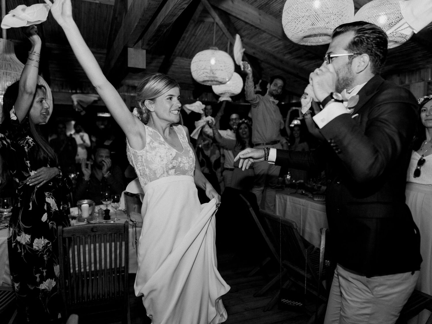 Photo en noir et blanc d'une mariée dansant joyeusement à la réception de mariage tandis que le marié boit à proximité, entourée d'invités.