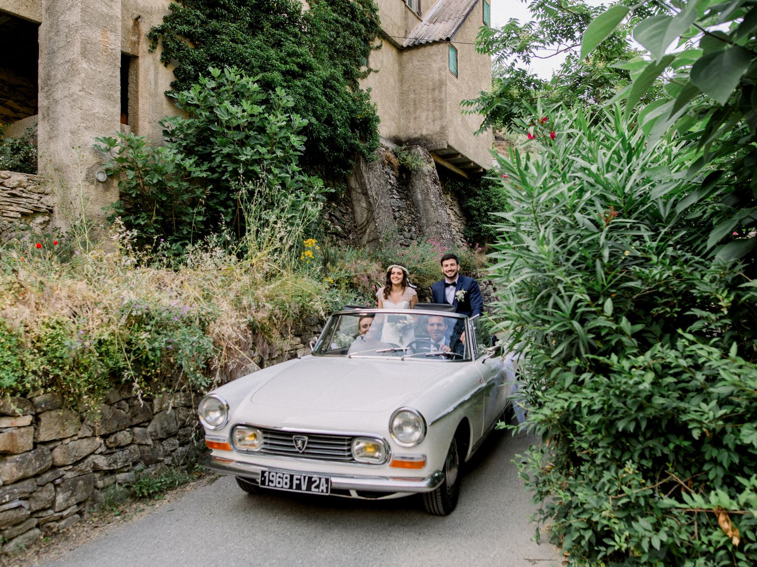 Bride and groom posing in a vintage white Peugeot convertible on a narrow lane surrounded by lush greenery.