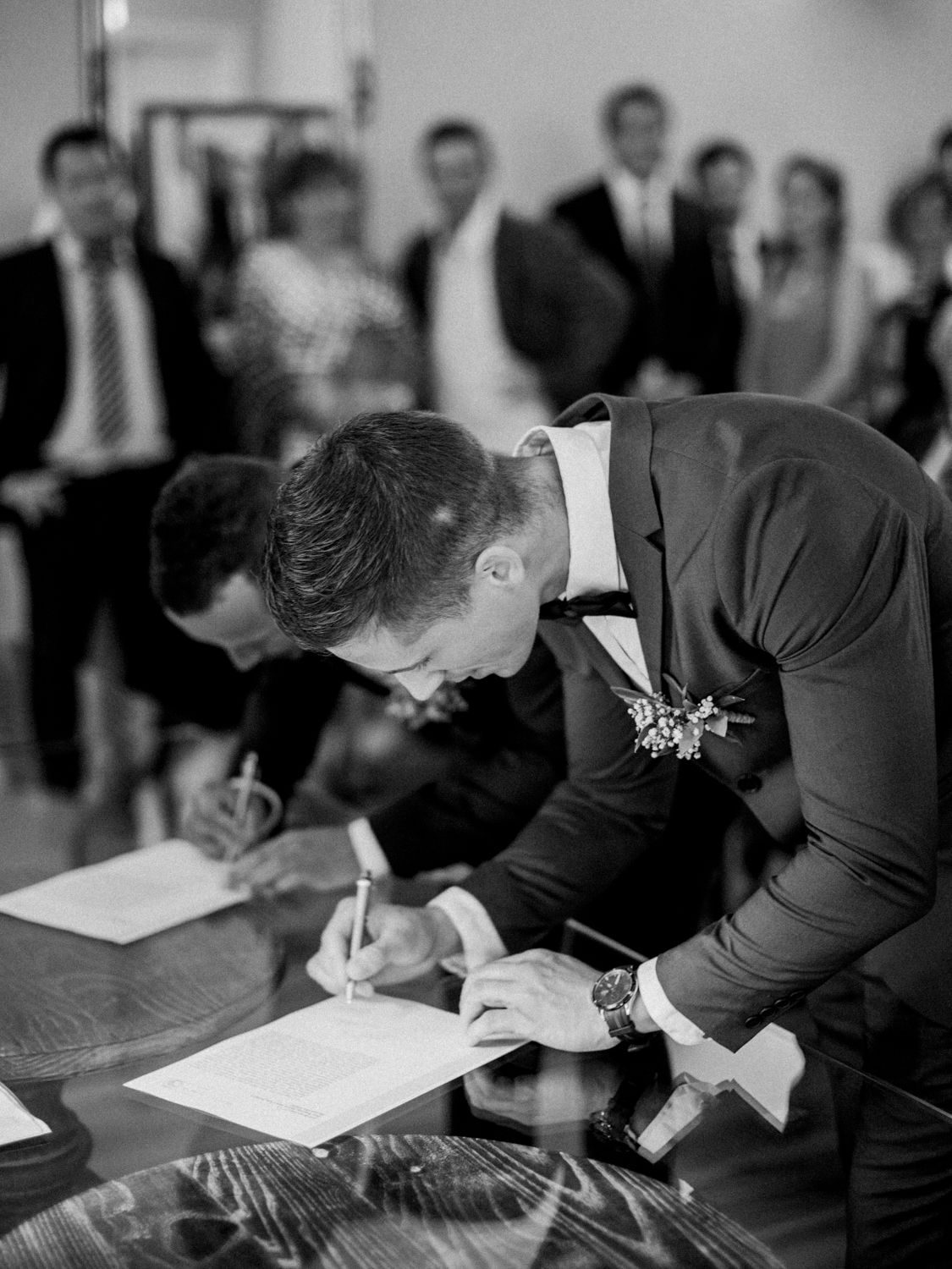 Groom signing wedding documents at ceremony, wearing suit with boutonniere and watch, guests in background, black and white.