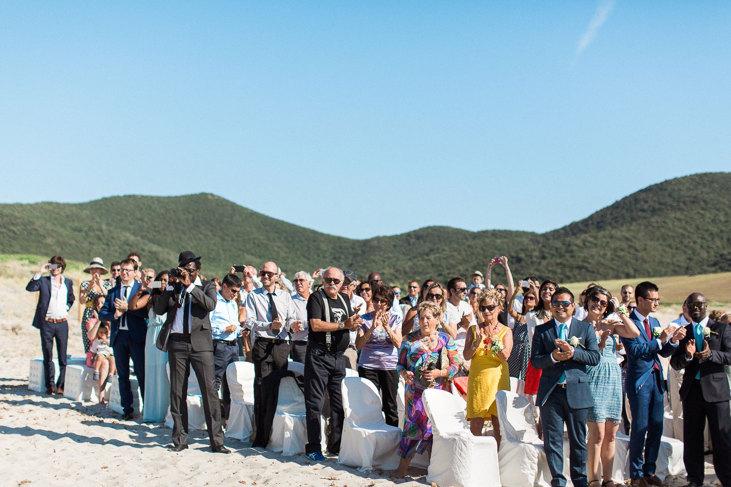 Des invités célèbrent un mariage en plein air sur une plage, avec des collines verdoyantes et un ciel bleu en arrière-plan.