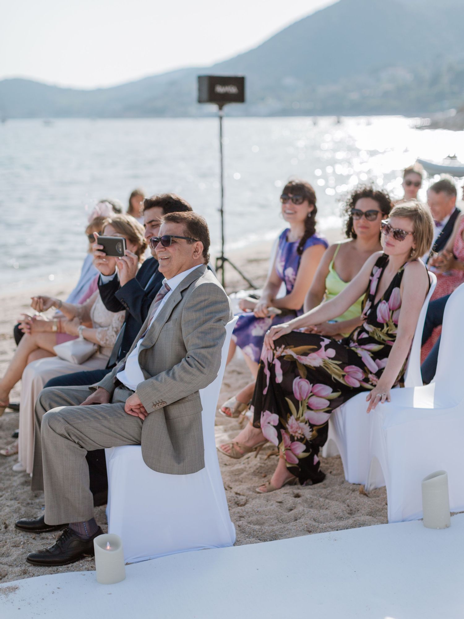 Des invités portant des lunettes de soleil, assis sur des chaises blanches lors d'une cérémonie de mariage sur la plage avec vue