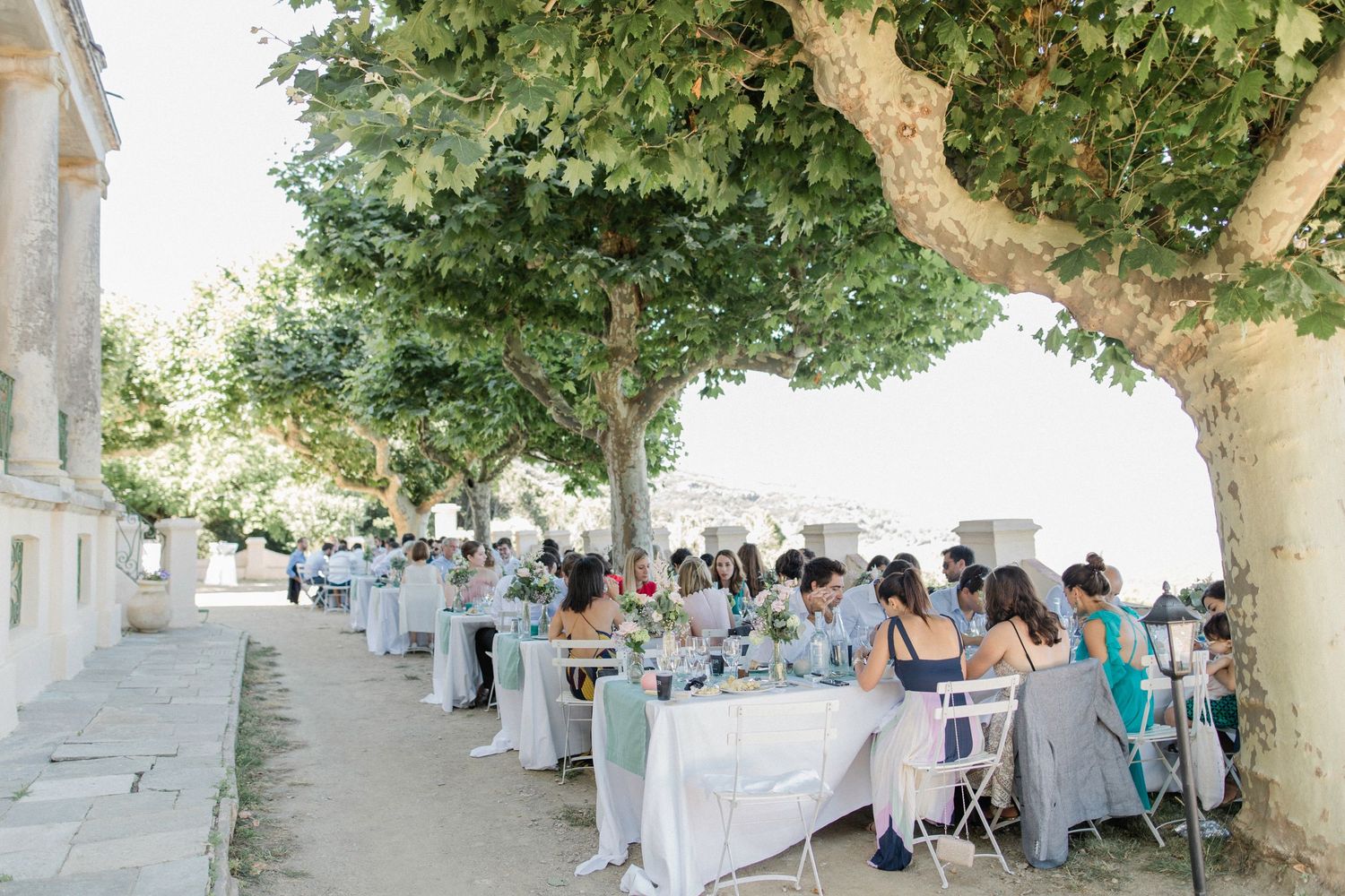 Outdoor wedding reception under lush green trees with guests dining at long white-draped tables in a Mediterranean setting.