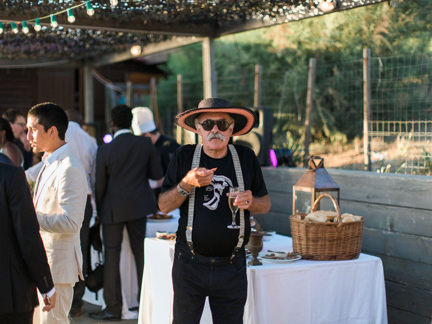 Un homme portant un sombrero et des lunettes de soleil tient un verre lors d'une fête en plein air avec des invités et des tables blanches.