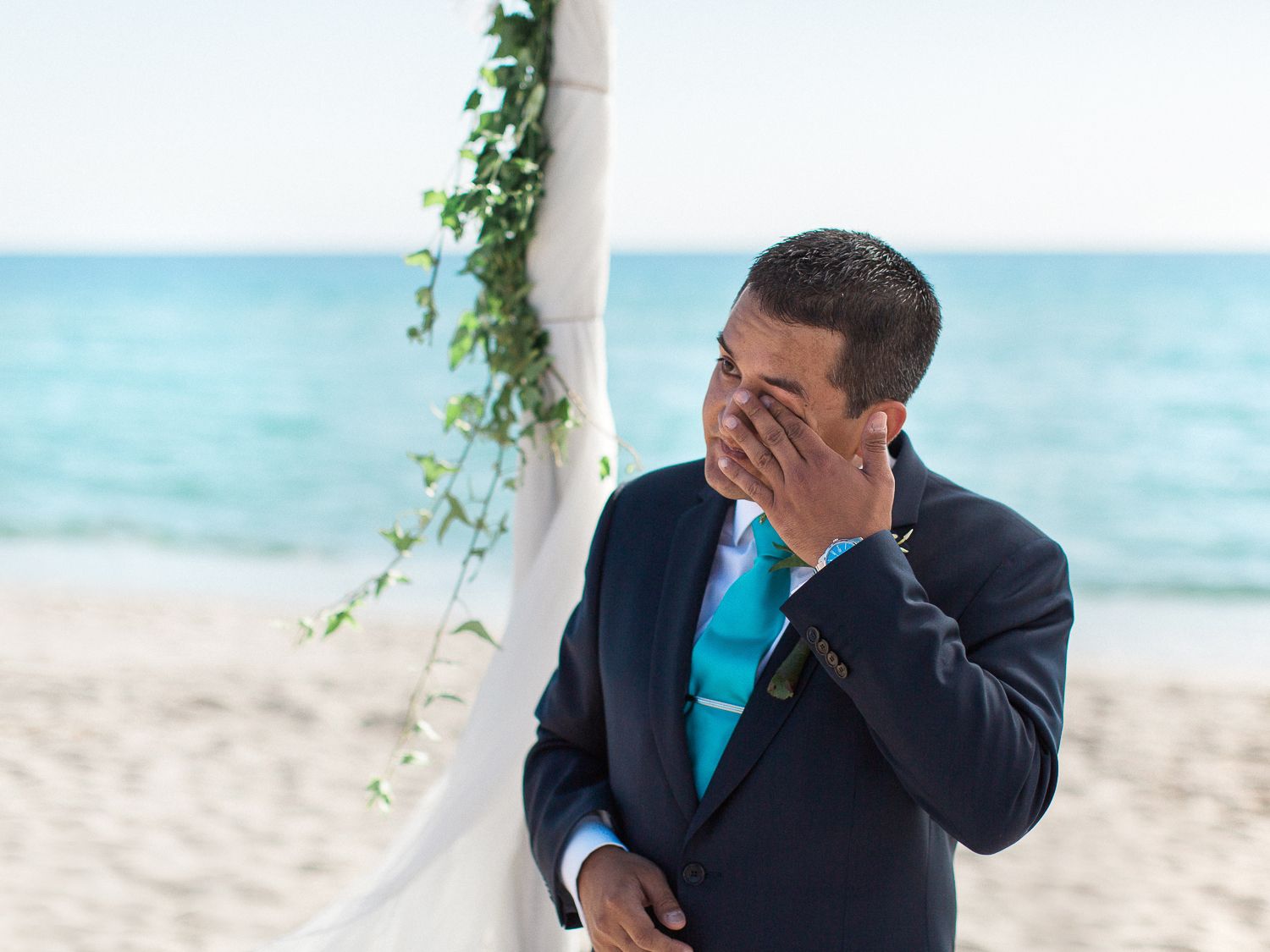 Un marié ému, en costume bleu marine, essuie ses larmes lors d'une cérémonie de mariage sur la plage, avec une arche blanche