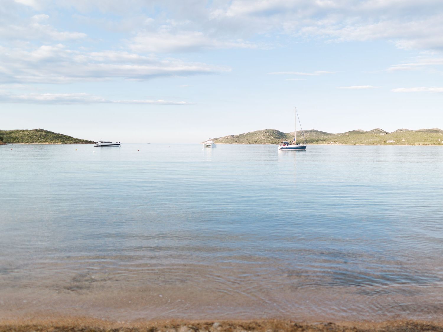 Baie côtière calme avec des voiliers ancrés sur une eau bleue limpide, des collines verdoyantes et un rivage sablonneux sous un ciel nuageux