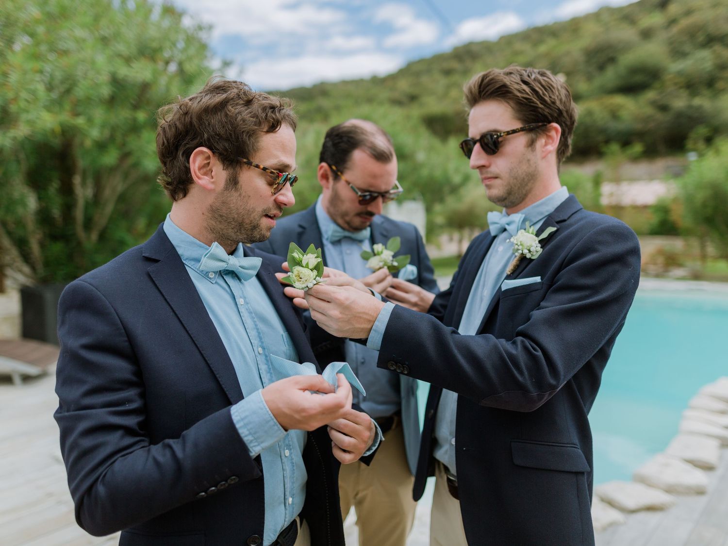 Trois garçons d'honneur en costumes bleu marine et nœuds papillon épinglent des boutonnières près d'une piscine lors d'un mariage