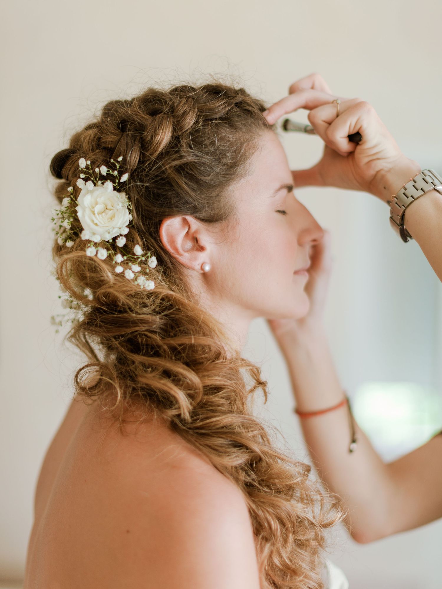Une mariée se fait maquiller ; elle porte des roses blanches et des fleurs de gypsophile dans ses cheveux bruns bouclés.