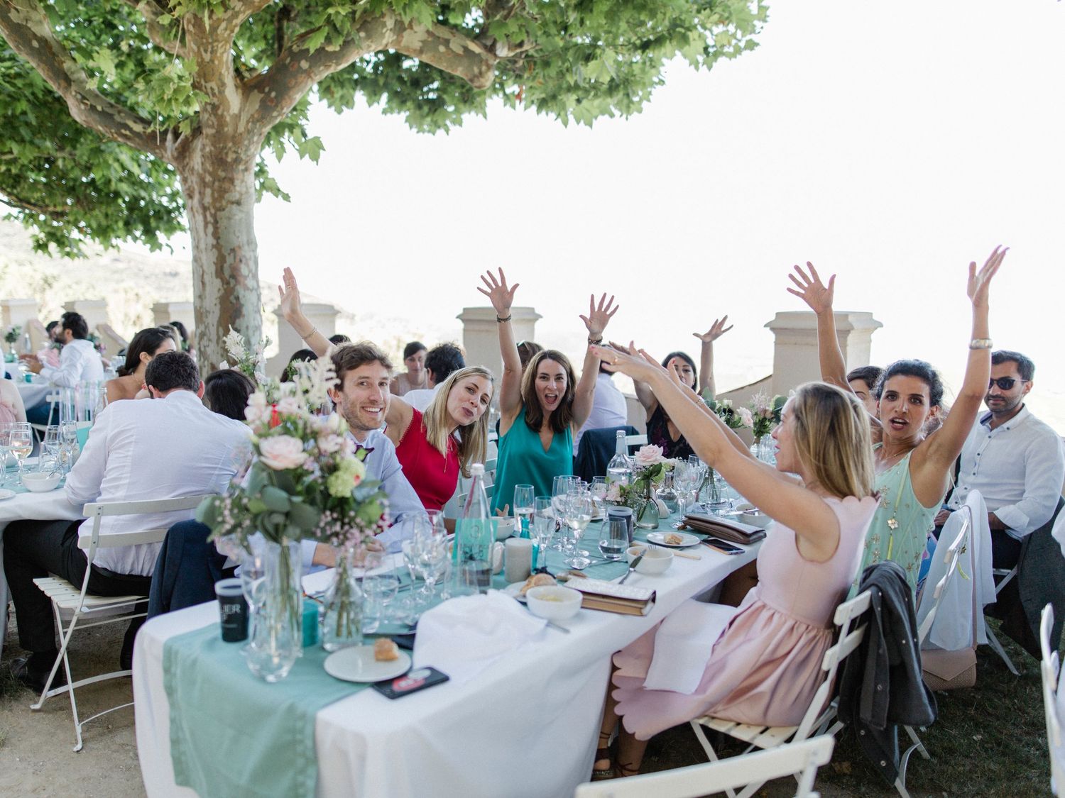 Guests celebrating at an outdoor wedding reception table under trees, raising hands joyfully with floral centerpieces.