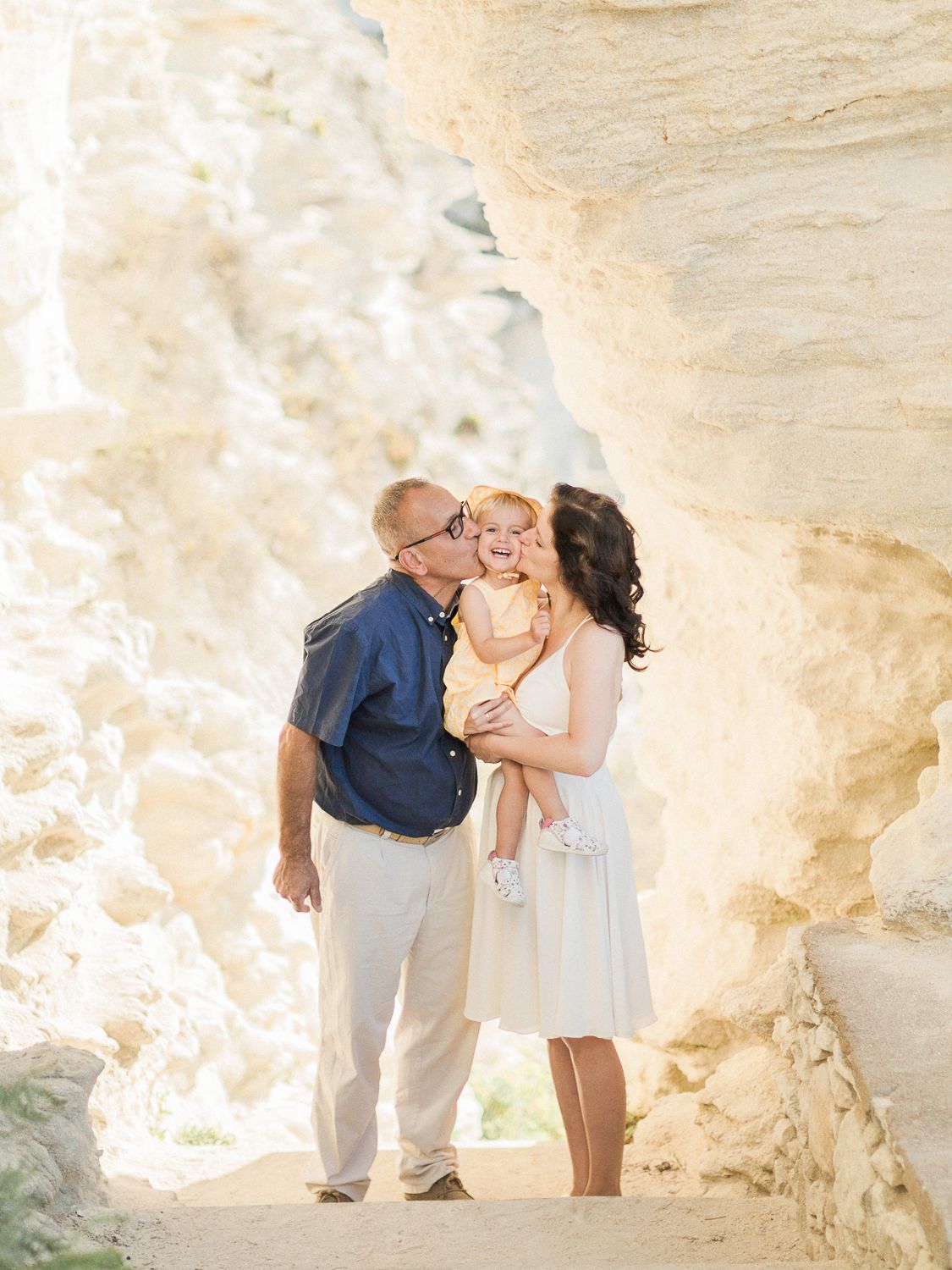 Des parents embrassent leur tout-petit rieur entre des formations rocheuses de grès doré, en plein air à Bonifacio