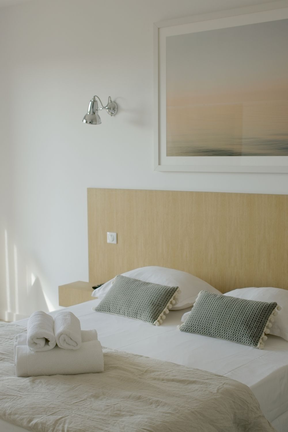 Chambre d'hôtel minimaliste avec tête de lit en bois, oreillers texturés verts, serviettes pliées et œuvre d'art côtière au-dessus du lit.