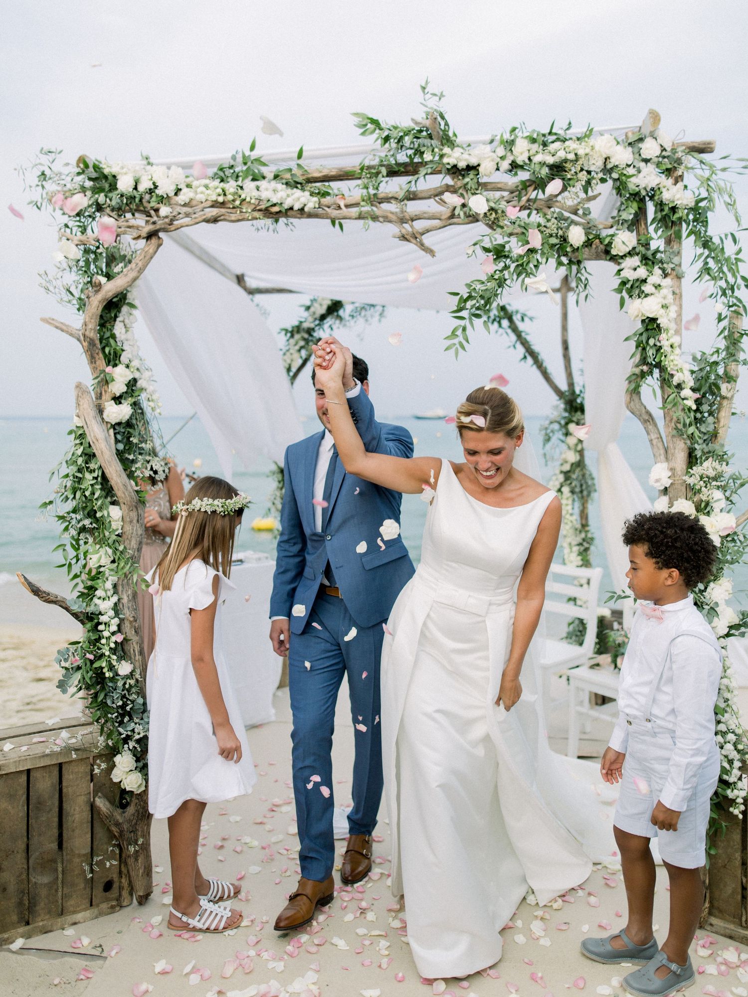Newlyweds celebrate under a floral beach arch with flower girl and ring bearer tossing rose petals.