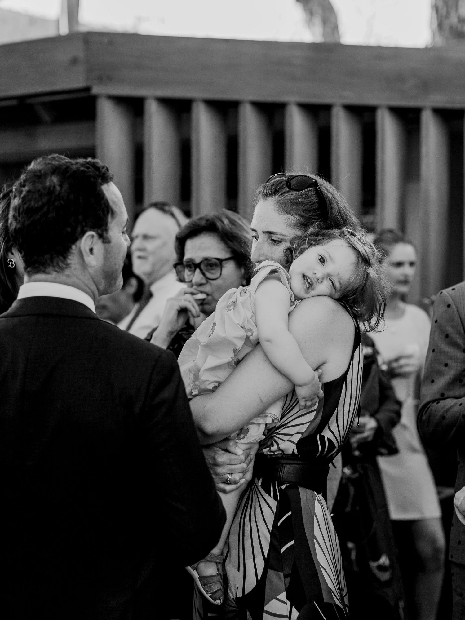 Photo en noir et blanc d'une femme en robe à motifs floraux tenant un enfant somnolent lors d'un événement en plein air.