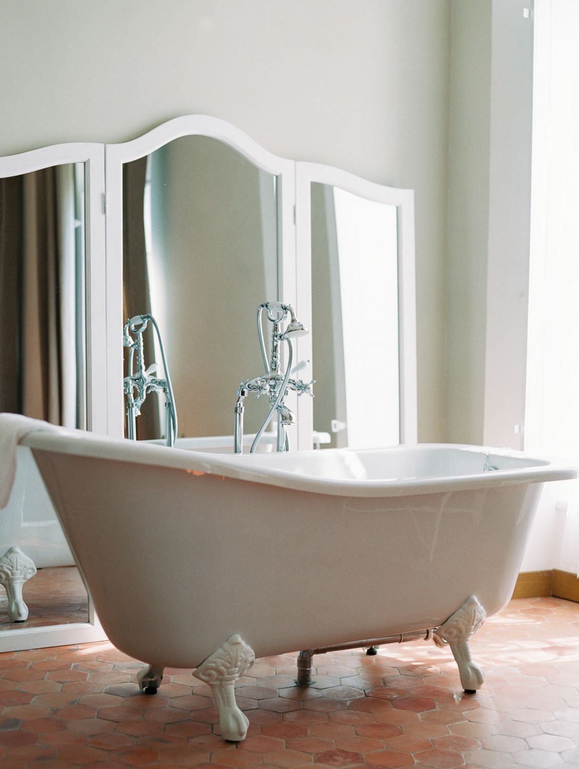 Elegant white clawfoot bathtub with chrome faucets on terracotta tile floor, reflected in a vintage arched mirror.