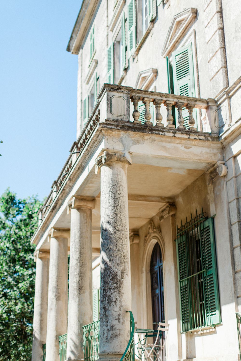 Historic European villa with green shutters, ornate balustrade balcony, and classical columns under a clear blue sky.