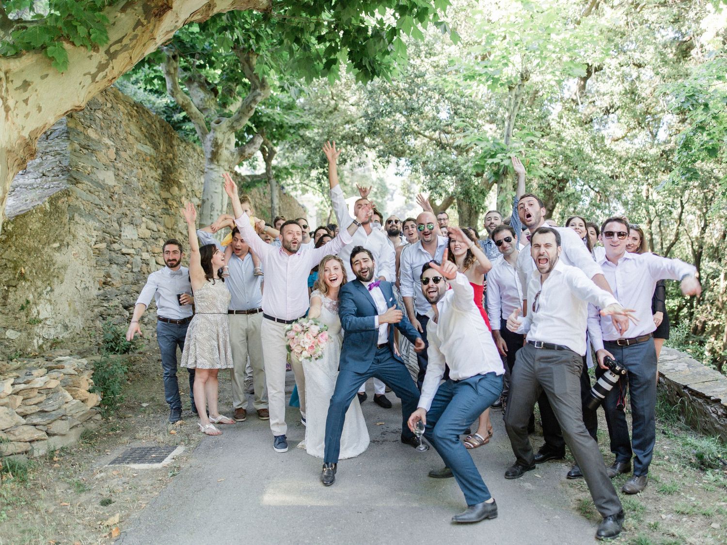 Joyful wedding group photo outdoors with bride, groom, and guests celebrating under lush green trees near a stone wall.