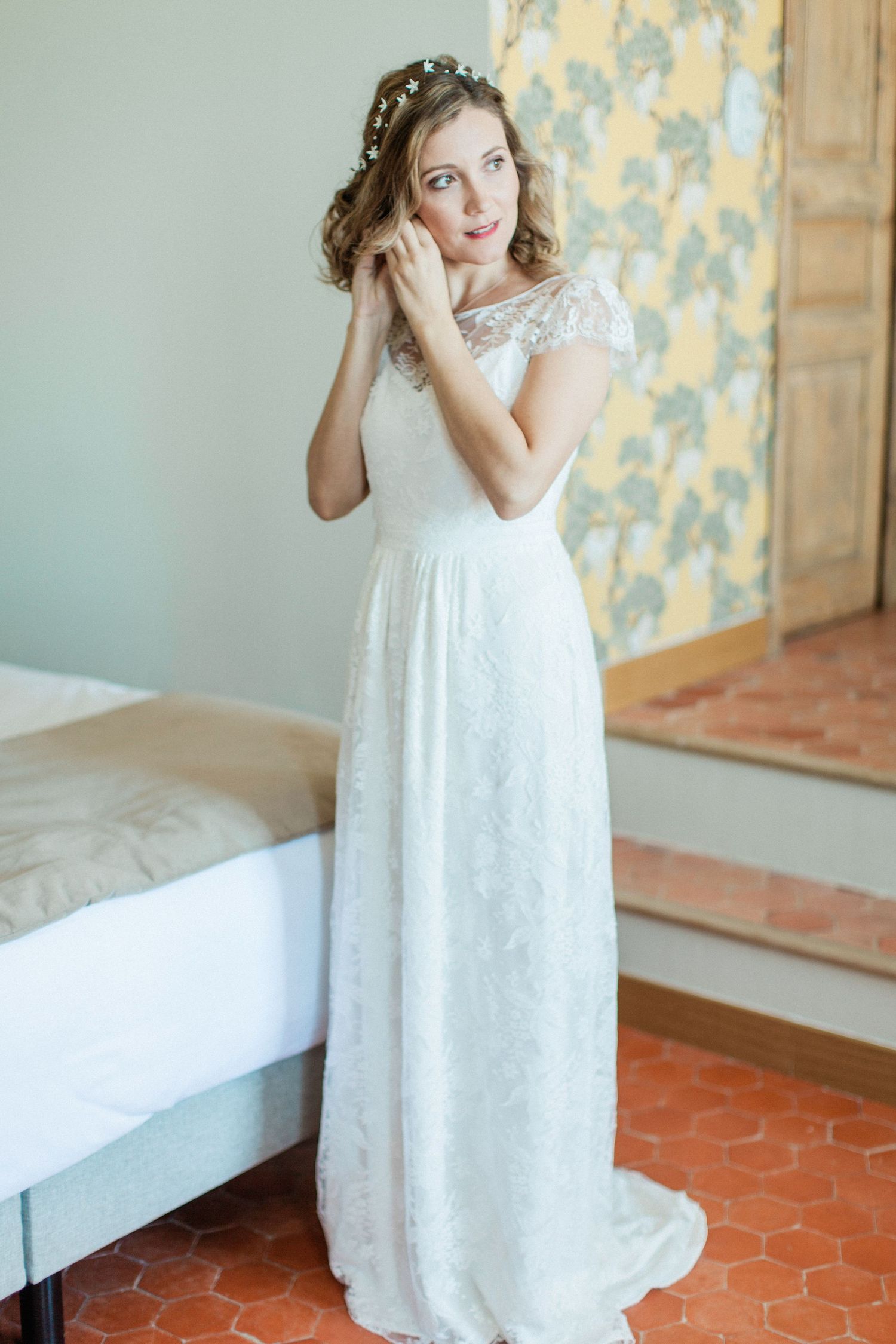 Elegant bride in white lace gown adjusting earring, wearing floral crown, standing in a sunlit room.